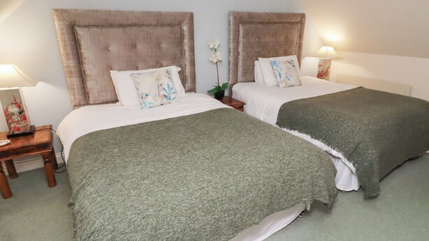 A hotel room with two beds, each with a headboard, a nightstand, a lamp, and a floral decoration. - Grange Cottage (Belford)