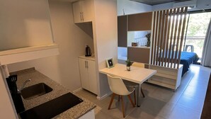 A compact kitchen with a granite countertop, white cabinets, and a built-in sink. There is a dining area with a white table and chairs, and a view through a window with a wooden blind. - Back to the Neighborhood - Luminous Studio in South Neighborhood with Pool (San Miguel de Tucumán)