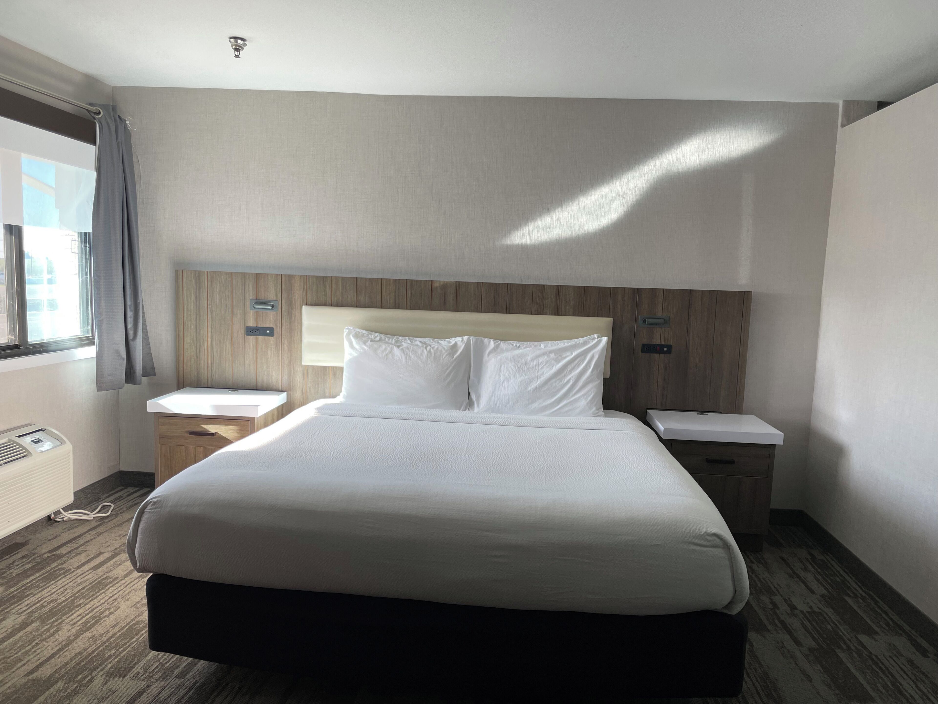 A hotel room with a large bed, wooden headboard, two bedside tables, a window with curtains, and a wall-mounted thermostat.