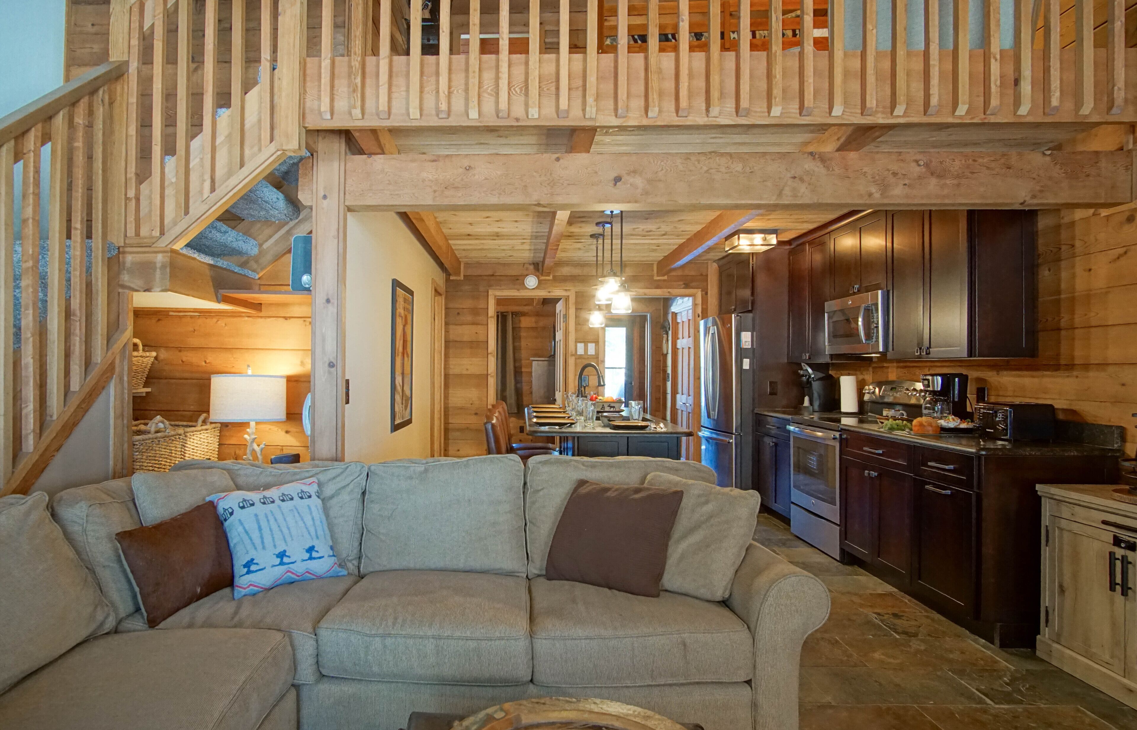 Ein gemütliches Wohnzimmer mit einer Holztreppe, einer Küche mit dunklen Schränken und Edelstahlgeräten sowie einem bequemen Sofa mit Kissen.