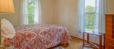 Un dormitorio con cama, mesita de noche, lámpara, cómoda y dos ventanas con cortinas.