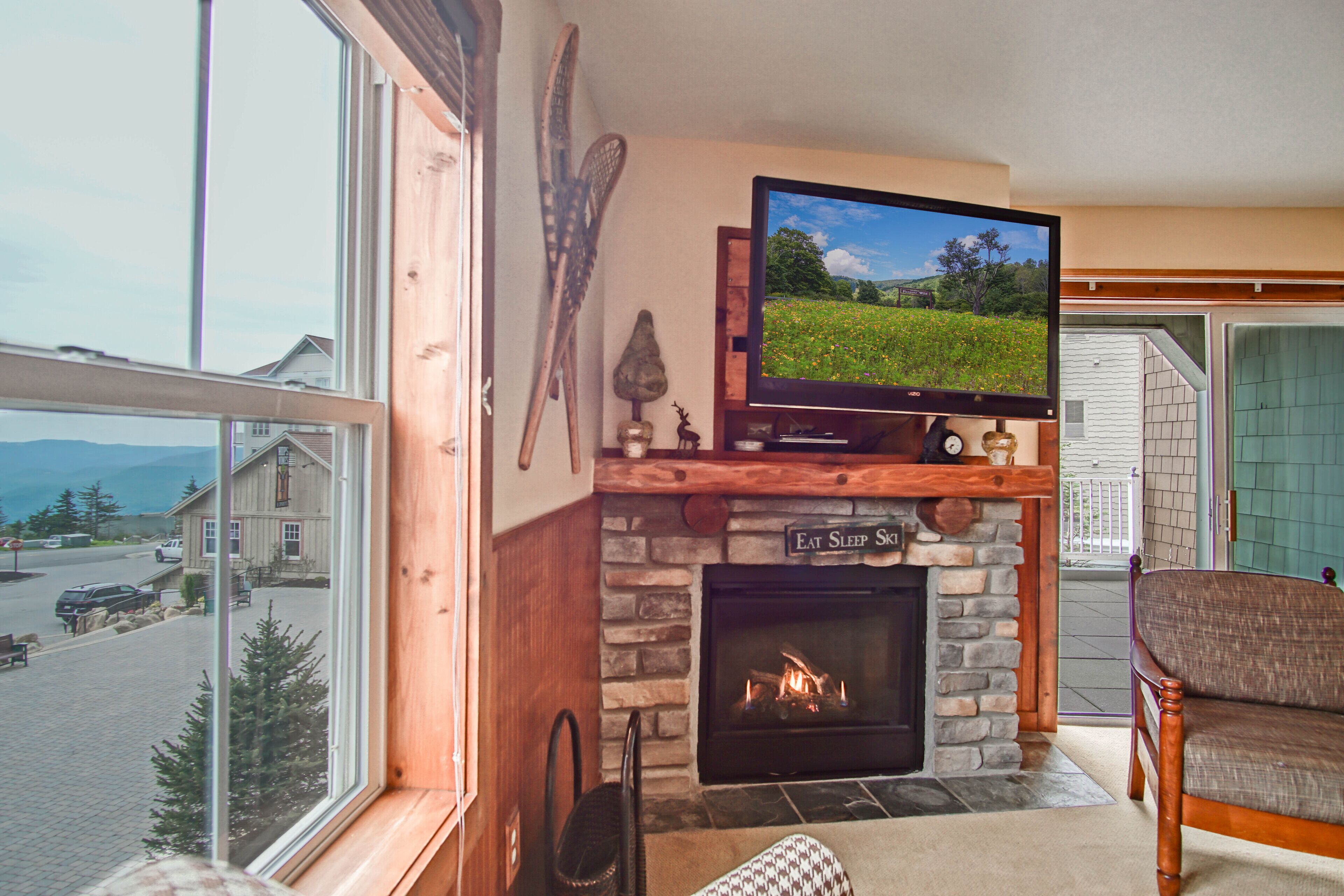 A cozy living room with a fireplace, a television mounted above it, and a view of the outside through large windows.