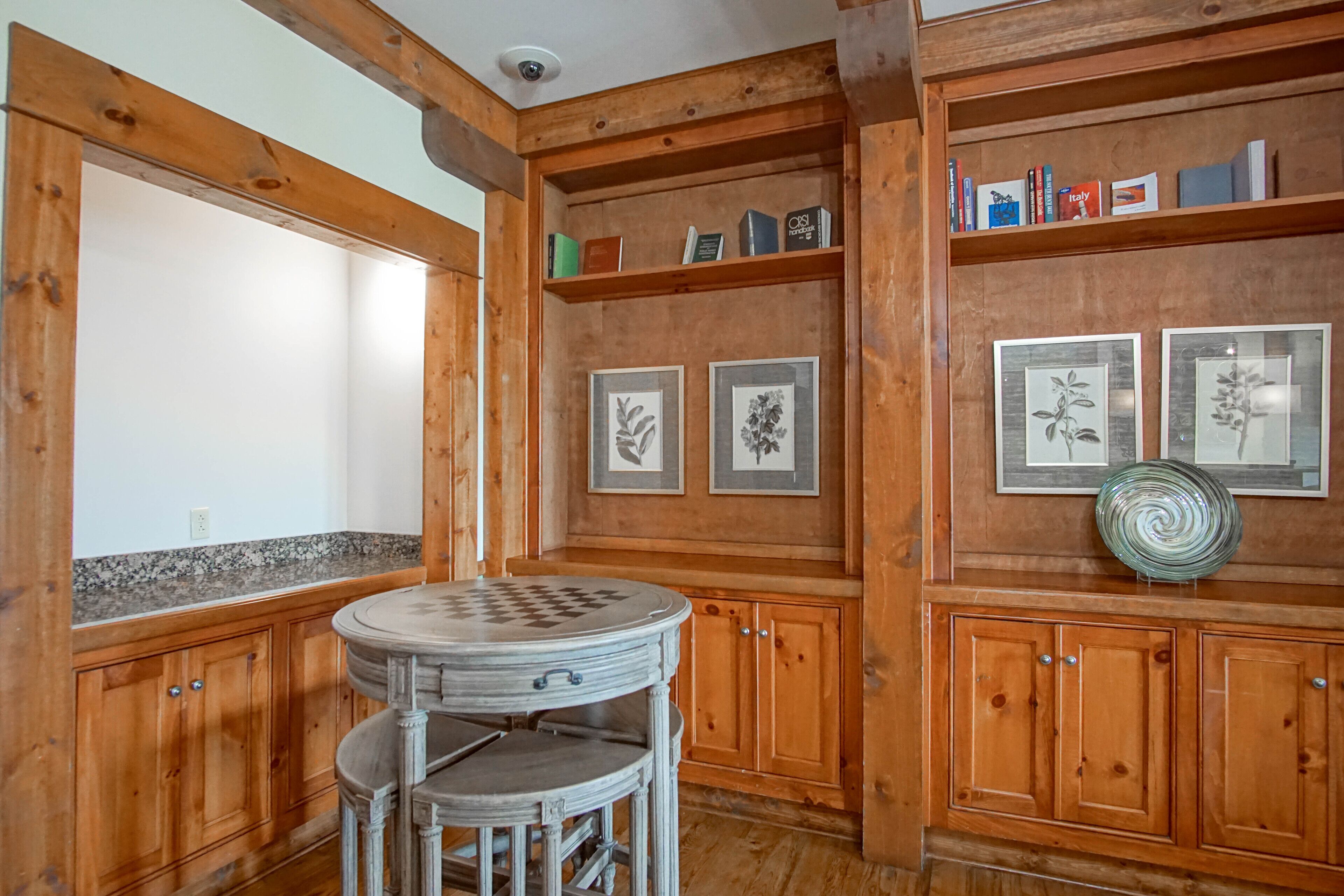 A wooden interior with a round table, framed artwork, and bookshelves.