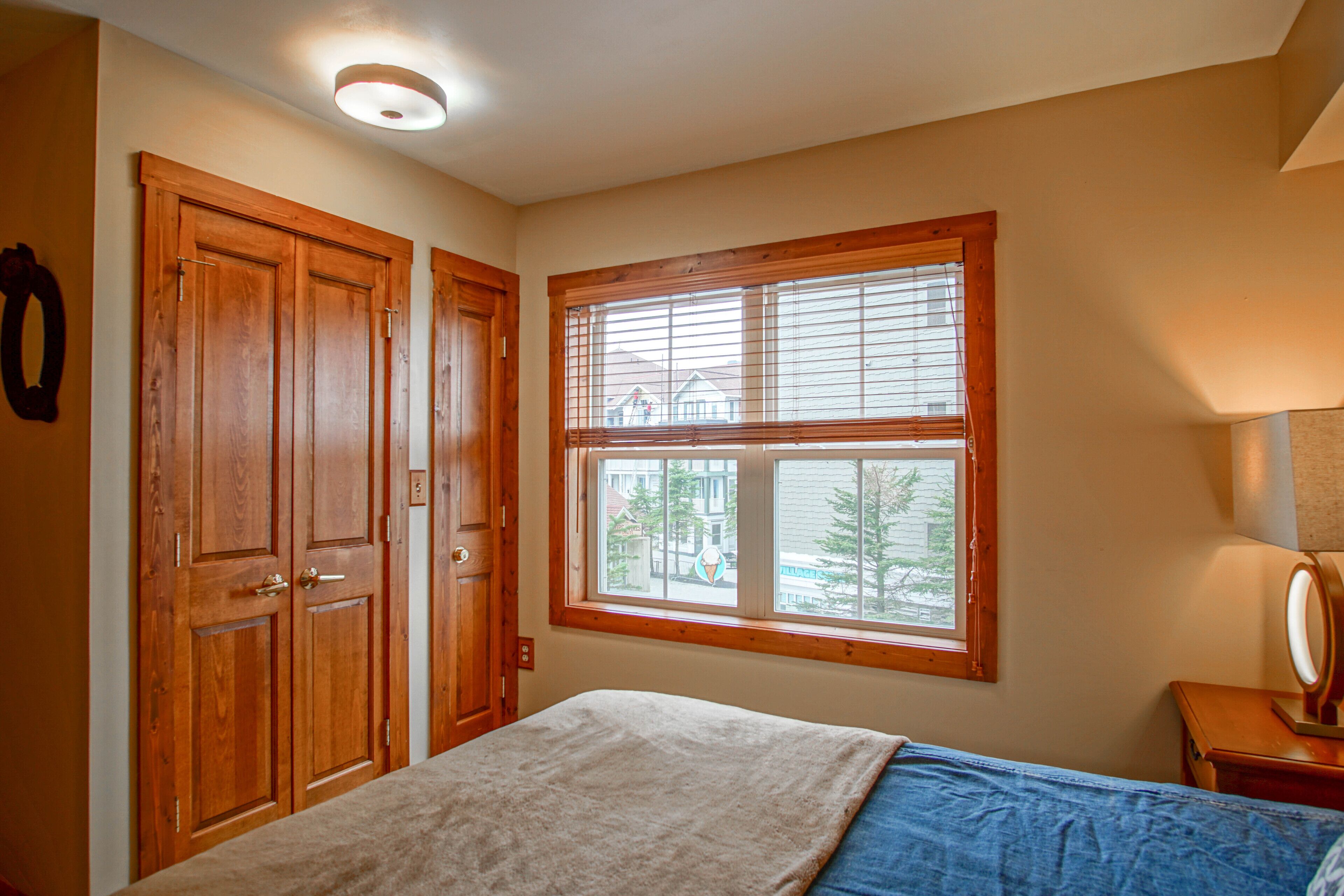 Un dormitorio con cama, ventana con persianas, puertas de madera y mesita de noche con lámpara.