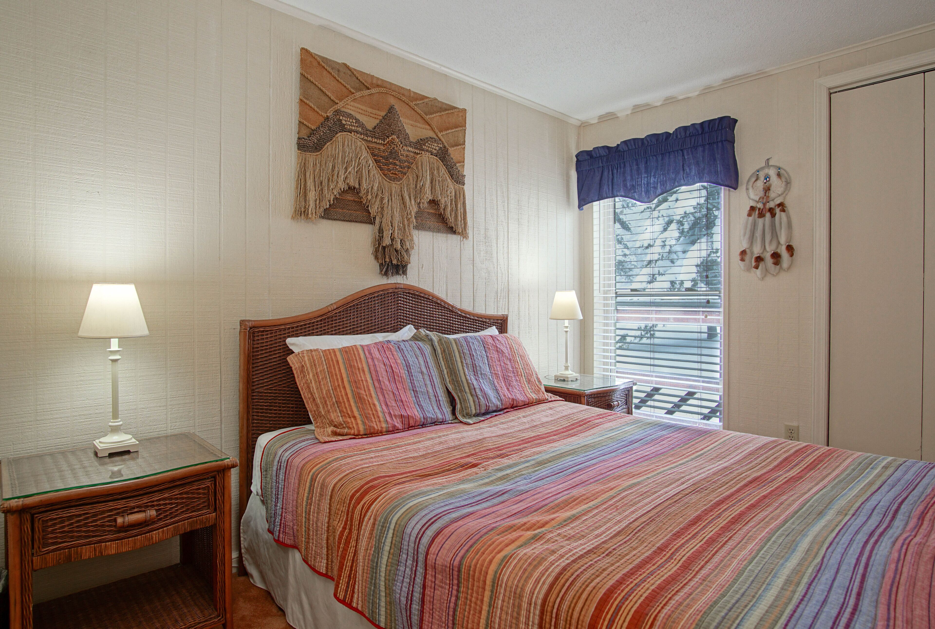 A bedroom with a large bed, two bedside tables, a wall hanging, and a window with blinds.
