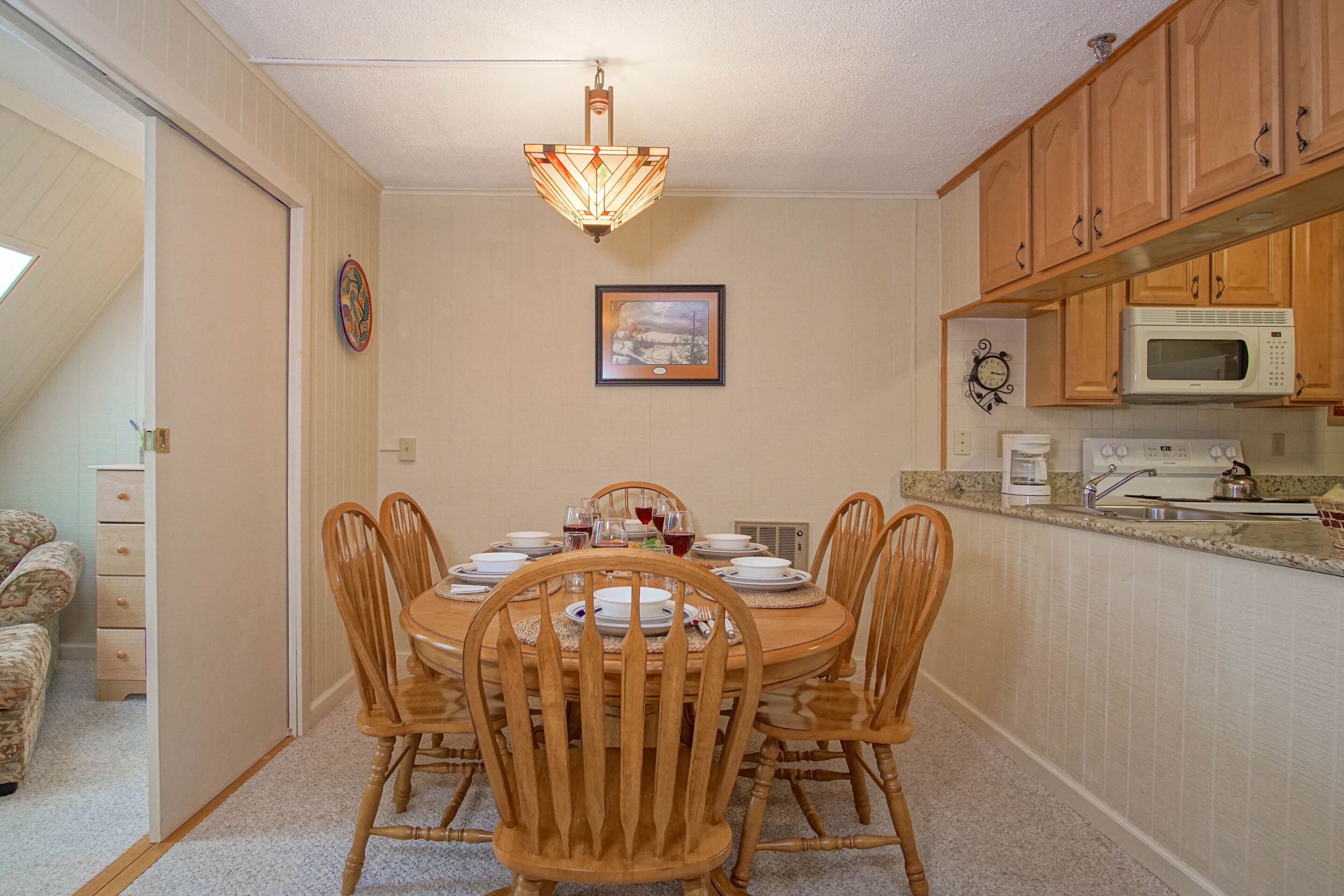 A dining room with a wooden table set for four, surrounded by chairs. There is a chandelier above the table, and a framed picture hangs on the wall.