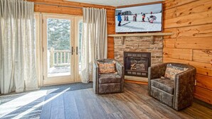 Una habitación acogedora con chimenea, dos sillones de cuero, un ventanal con cortinas y un televisor mostrando un paisaje nevado.