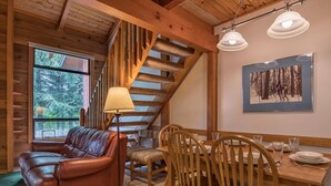 Interior acogedor de una cabaña con sofá de cuero, mesa de comedor de madera, sillas, una lámpara, una escalera y un cuadro en la pared.