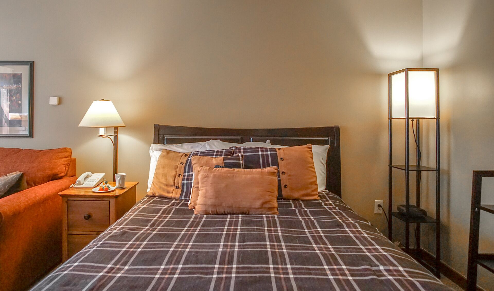 A hotel room with a bed, a nightstand, a lamp, a telephone, and a framed picture on the wall.