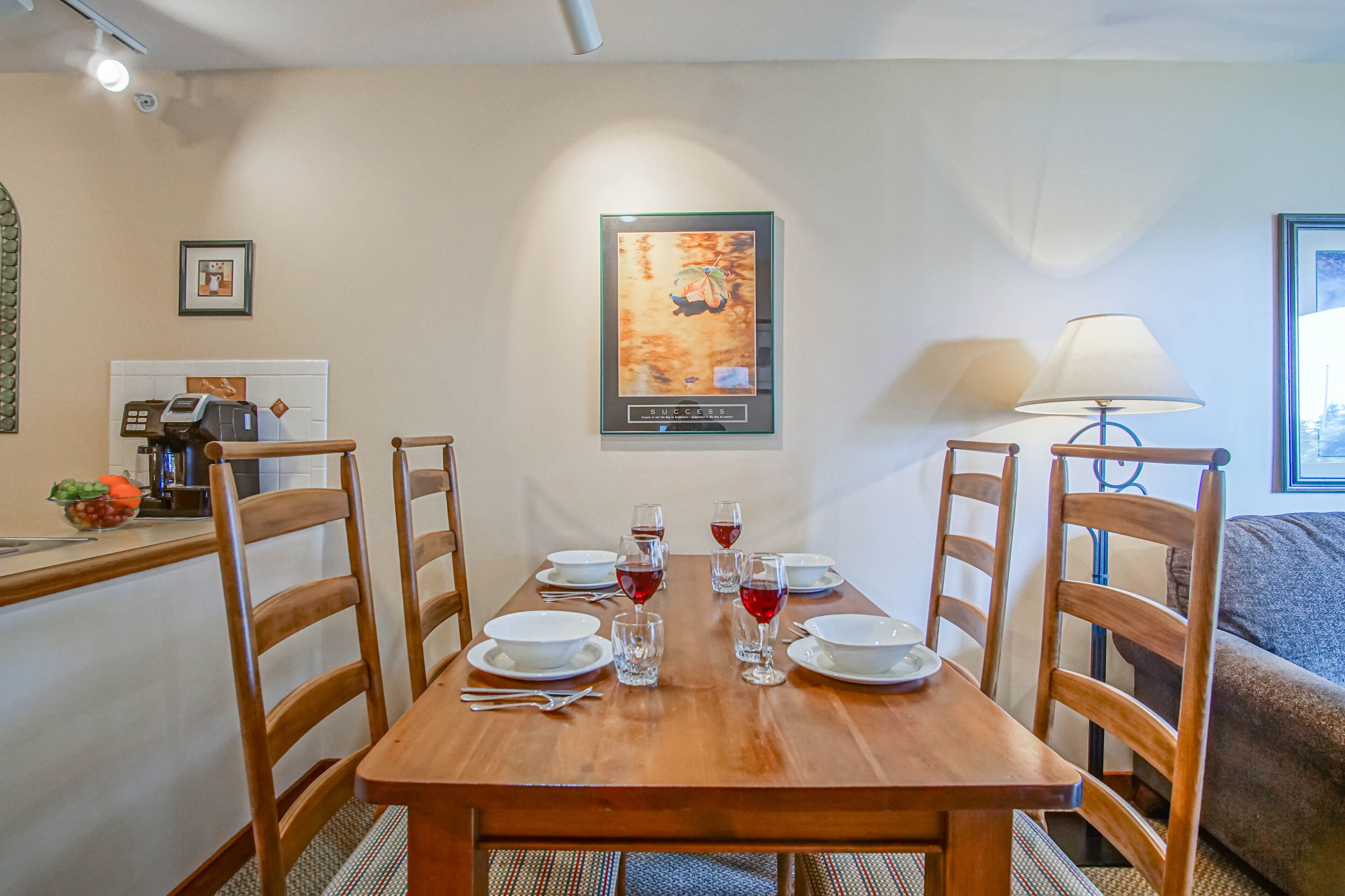A dining area with a wooden table set for four, surrounded by wooden chairs. There are plates, glasses, and cutlery on the table. A framed picture hangs on the wall above the table.