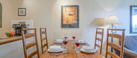 A dining area with a wooden table set for four, surrounded by wooden chairs. There are plates, glasses, and cutlery on the table. A framed picture hangs on the wall above the table.