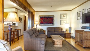 Ein gemütliches Wohnzimmer mit einer Couch, Sesseln, einem Couchtisch und einem Fernseher. Holzschränke und ein gemustertes Teppich sind ebenfalls vorhanden.