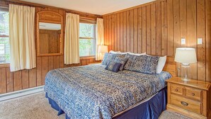 Ein Schlafzimmer mit einer holzverkleideten Wand, einem Bett mit gemusterten Betttextilien, einem Nachttisch mit Lampe und einem Fenster mit Vorhängen.