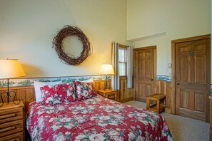 Un dormitorio con cubrecama floral, cabecera de madera, dos mesitas de noche con lámparas, una puerta de madera y una corona decorativa en la pared.