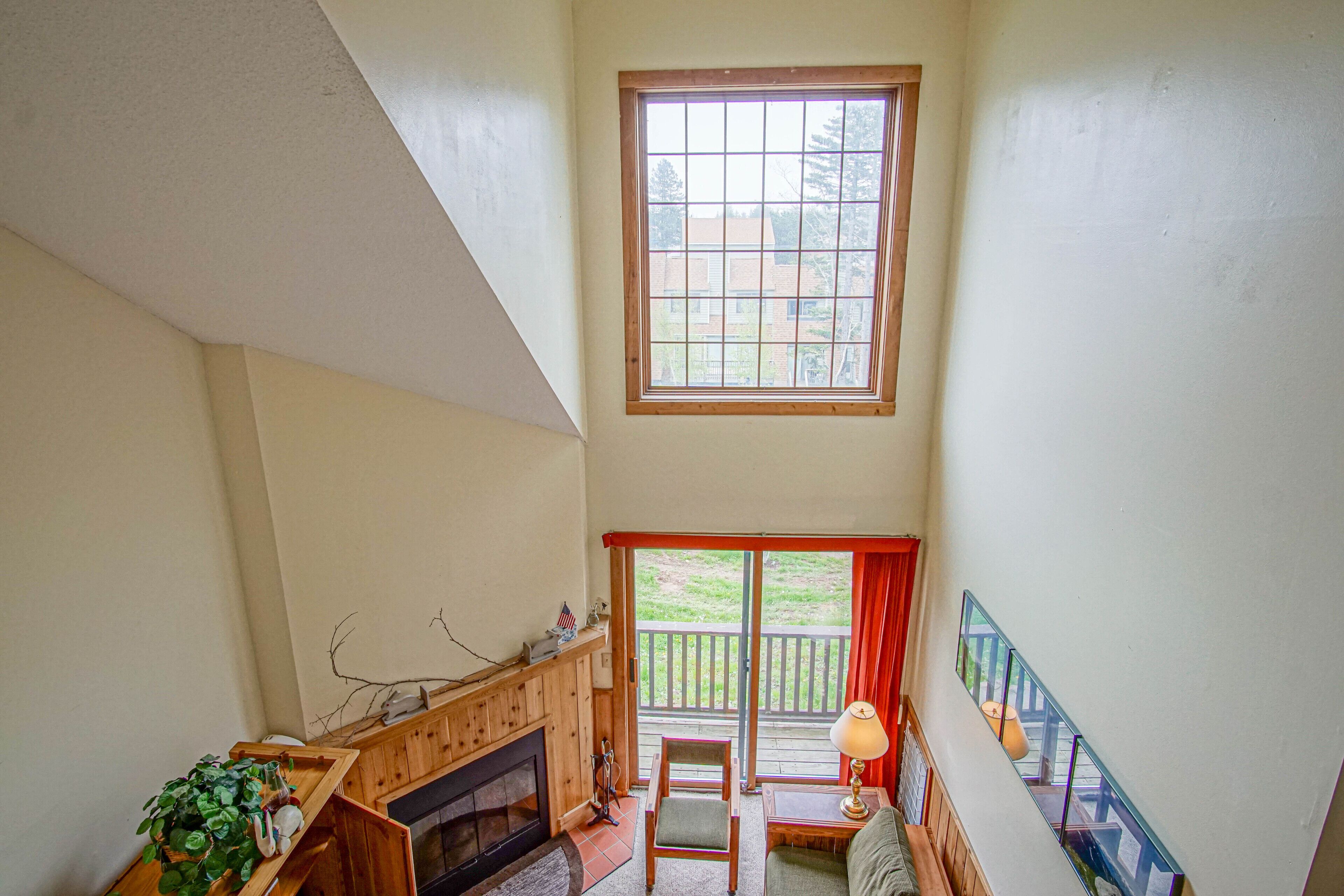 A high-ceilinged room with a fireplace, a window with a view of the outdoors, and a balcony with a railing.