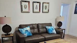 A living room with a brown leather sofa, two blue patterned throw pillows, a wooden coffee table, and two side tables with lamps. Three framed pictures are hung on the wall above the sofa. - 103 Classic Studio Suite in waterfront hotel in Singer Island, FL (Riviera Beach)