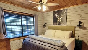 A bedroom with a bed, a ceiling fan, a window with blinds, and a wall-mounted deer painting. - Stargazer Cabin: Sauna, Rooftop Deck, Fire Pit, Indoor Wood Fireplace, Privacy (Blue Ridge)