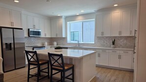 A modern kitchen with white cabinetry, a central island, two bar stools, a stainless steel refrigerator, and a window providing natural light. - Spacious 4-bedroom townhome in beautiful Long Beach with AC (Long Beach)