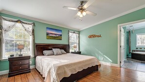 Une chambre avec un grand lit, un plancher en bois, un ventilateur de plafond et une fenêtre avec des rideaux.