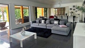 A modern living room with a grey sectional sofa, a white coffee table, and a black rug. There is a dining area with a white table and chairs, and a kitchen area with a white cabinet. Large windows provide views of the outdoors. - Villa Alana (Vaitape)