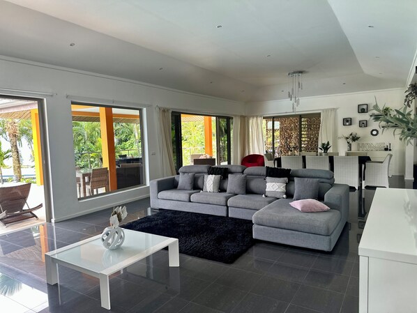 A modern living room with a grey sectional sofa, a white coffee table, and a black rug. There is a dining area with a white table and chairs, and a kitchen area with a white cabinet. Large windows provide views of the outdoors. - Villa Alana (Vaitape)