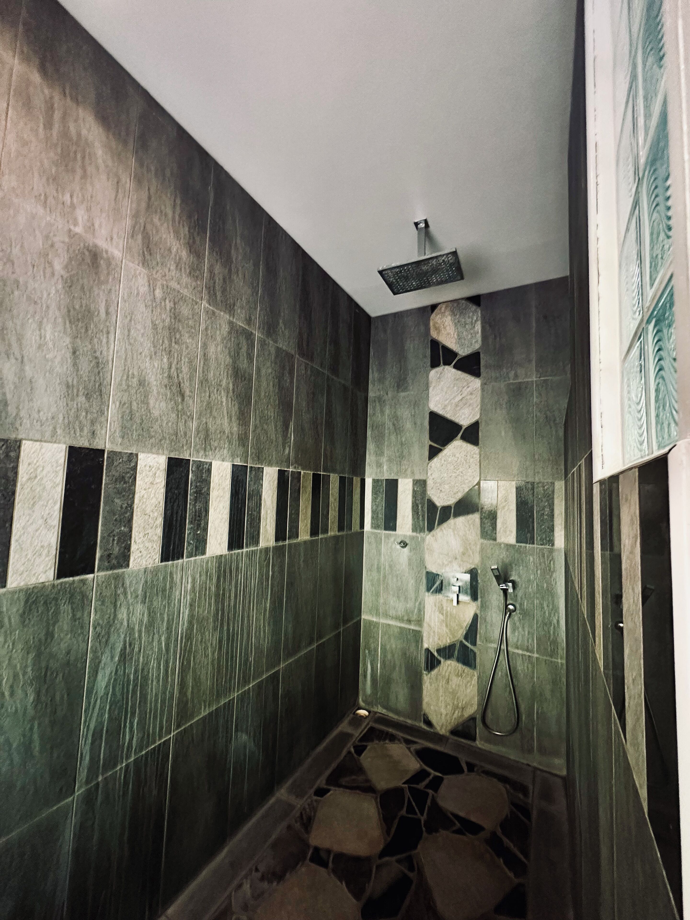 A bathroom with dark green tiles, a stone accent wall, and a shower area with a handheld showerhead.