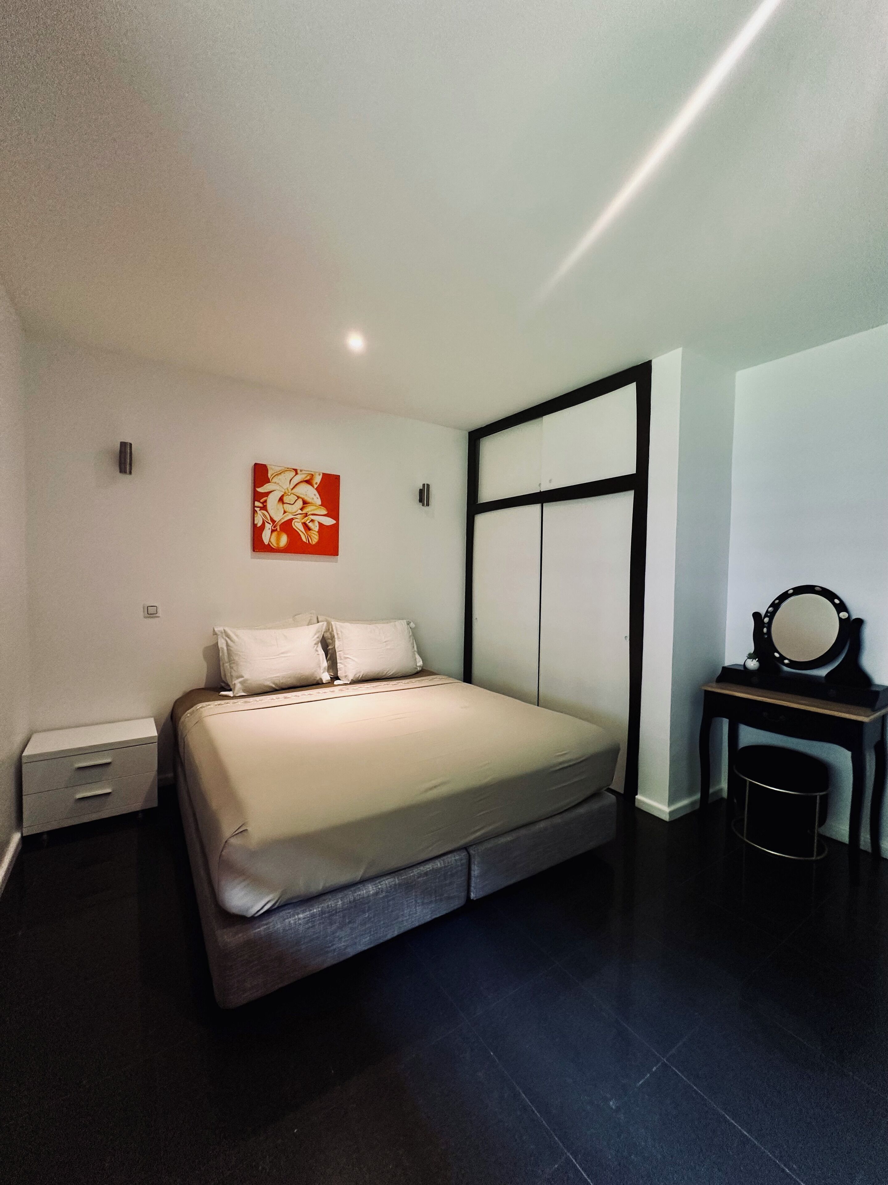 Une chambre à coucher moderne, avec un lit, une table de chevet, un bureau doté d’un miroir et une œuvre d’art accrochée au mur.