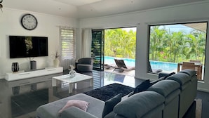 A modern living room with a flat-screen TV, a large sofa, a coffee table, and a view of a swimming pool and lush greenery. - Villa Alana (Vaitape)