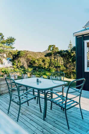 Una mesa de vidrio con sillas de metal en una terraza de madera, con vistas a un jardín exuberante y colinas.