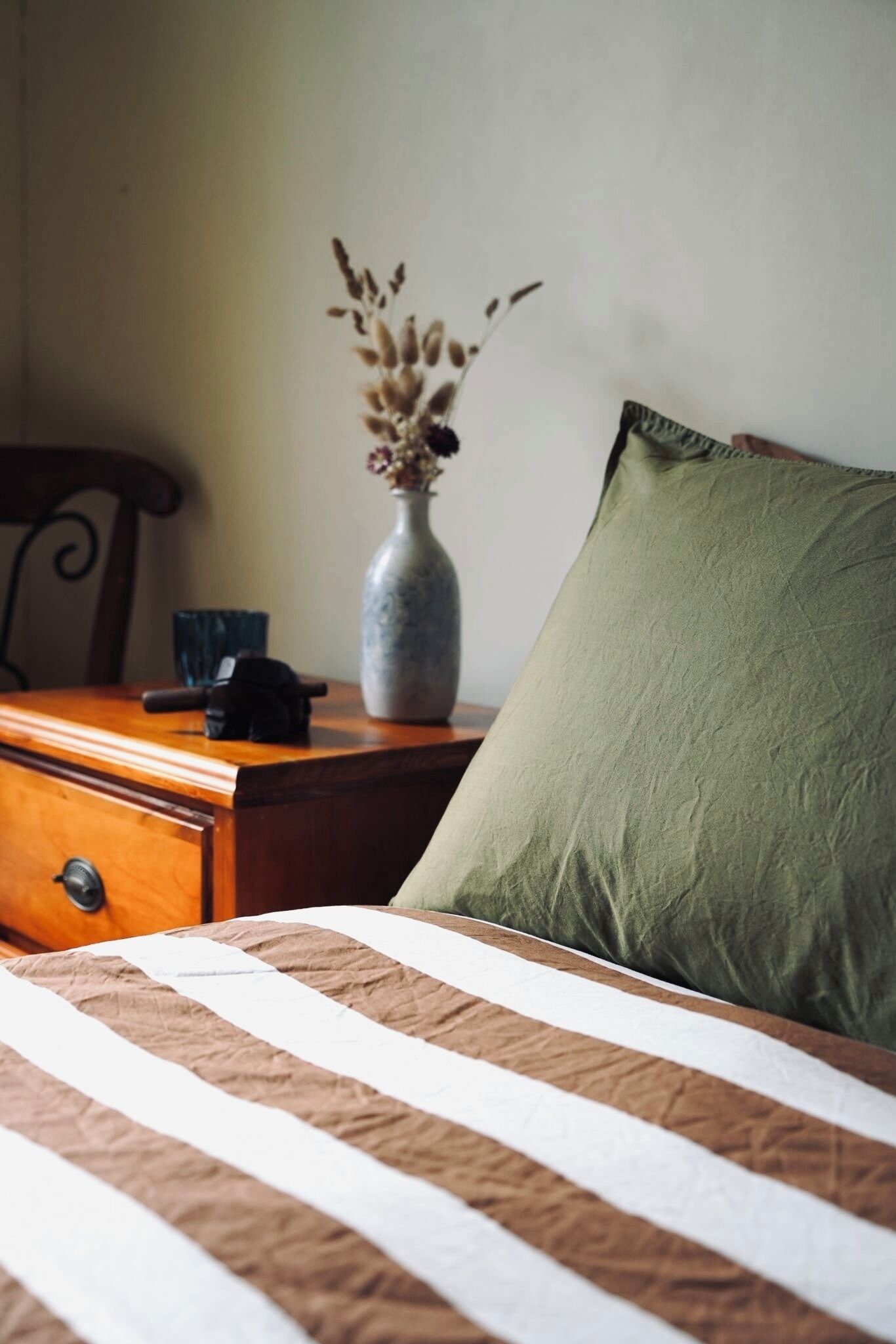Ein Bett mit gestreifter Bettdecke, ein grünes Kopfkissen, eine Vase mit getrockneten Blumen und ein Nachttisch aus Holz mit einem Telefon.