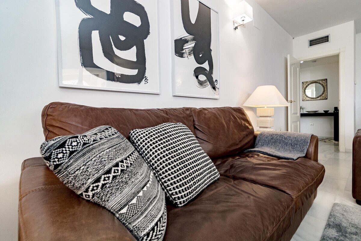 A brown leather sofa with patterned cushions, a wall-mounted lamp, and two large abstract paintings.