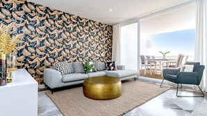 Ein modernes Wohnzimmer mit einem gemusterten Wandpapier, einem grauen Sofa mit gemusterten Kissen und einem runden Couchtisch aus Gold, mit Blick auf das Meer durch große Fenster.