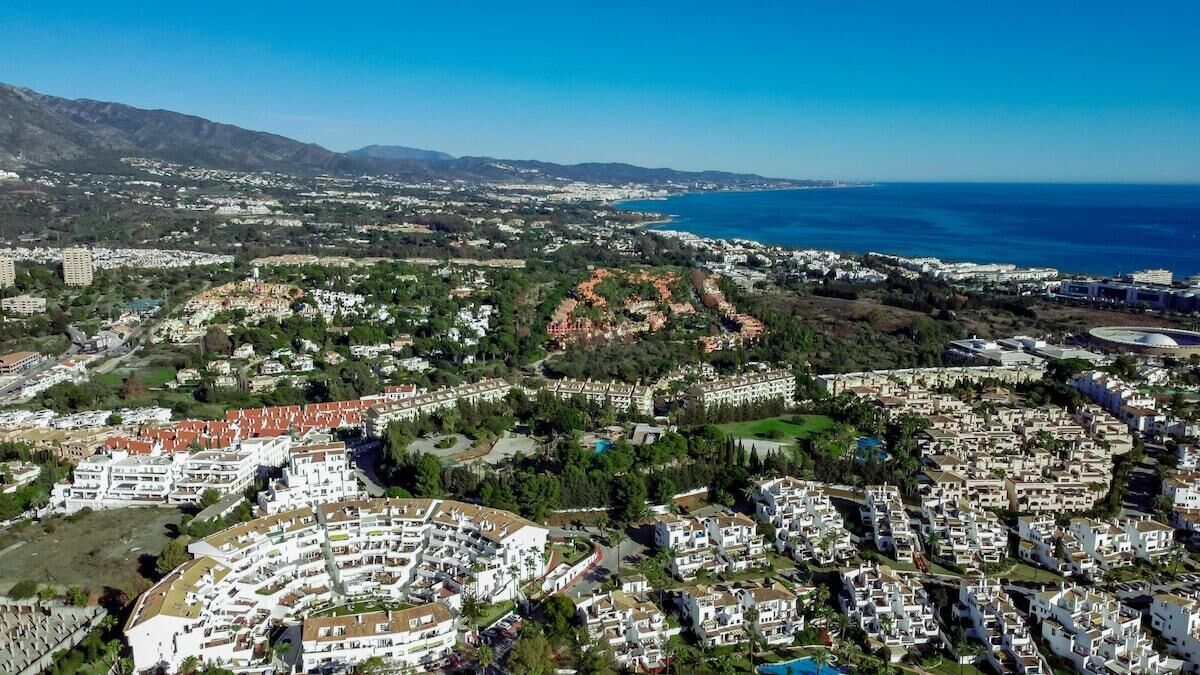 Vista aérea de un pueblo costero con edificios residenciales, un puerto deportivo y montañas al fondo.