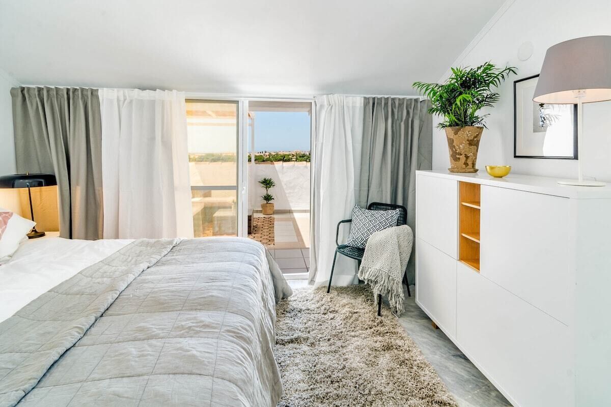 A bedroom with a bed, a chair, a desk, a plant, and a view of the outdoors.