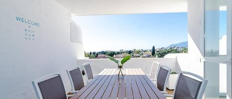 Een modern buitenontbijtgedeelte met een houten tafel en stoelen, een welkomstbord, en een uitzicht op de stad en de bergen op de achtergrond.