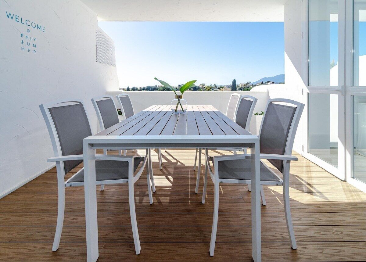 A white table with six chairs on a wooden deck, overlooking a scenic view.