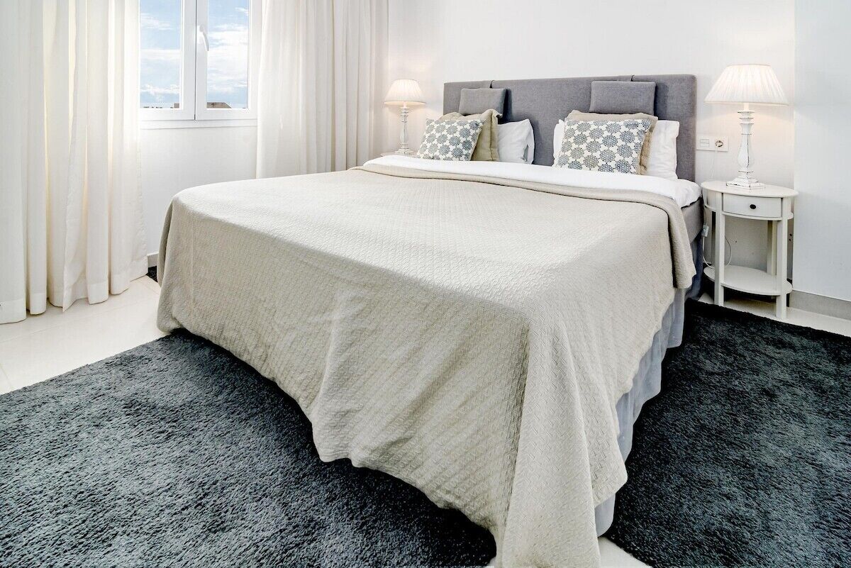 Una cama bien hecha con cabecera gris, ropa de cama blanca y almohadas estampadas. Hay dos mesitas de noche con lámparas. La habitación tiene un ventanal con cortinas blancas.