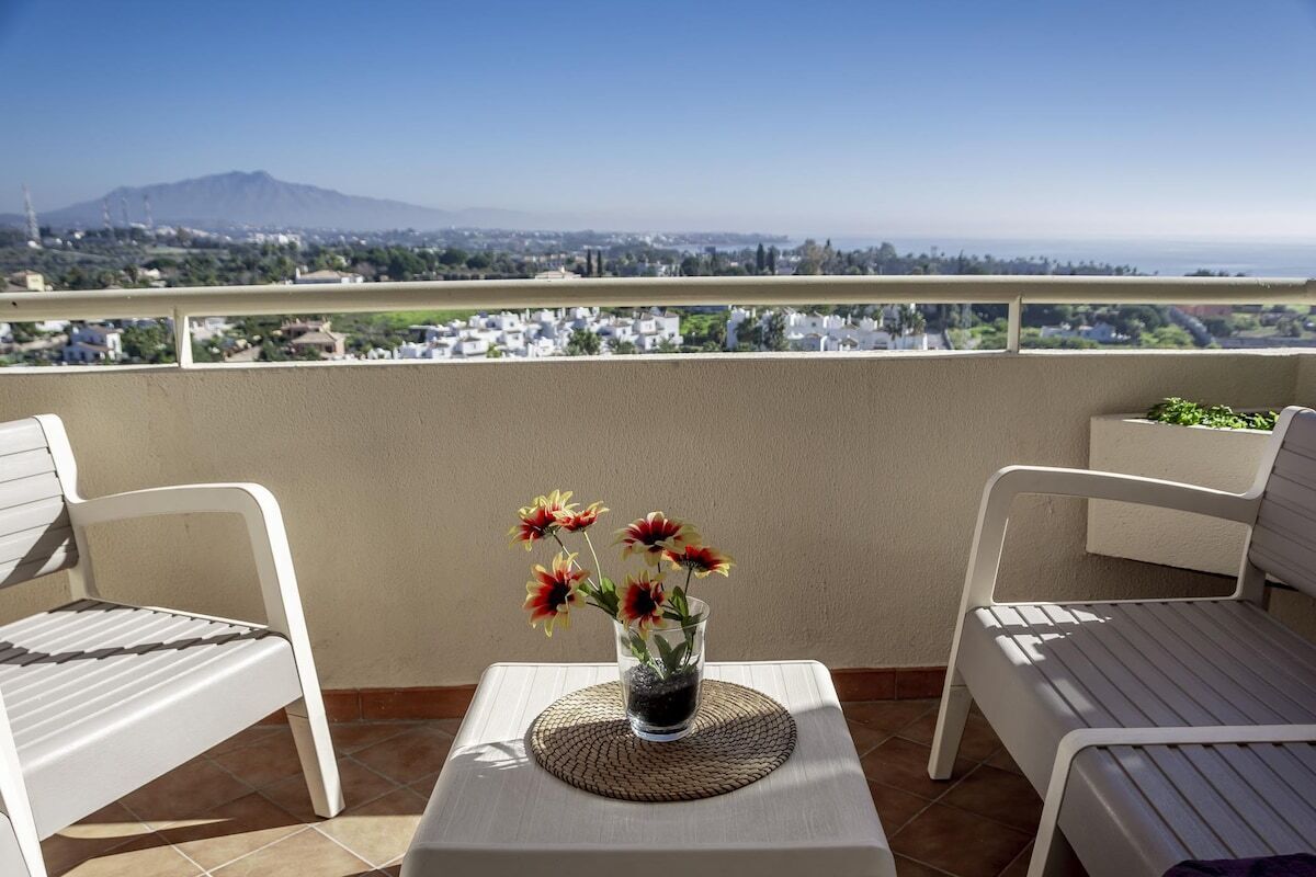 A balcony with two white chairs, a small table with a vase of flowers, and a view of a distant mountain and cityscape.