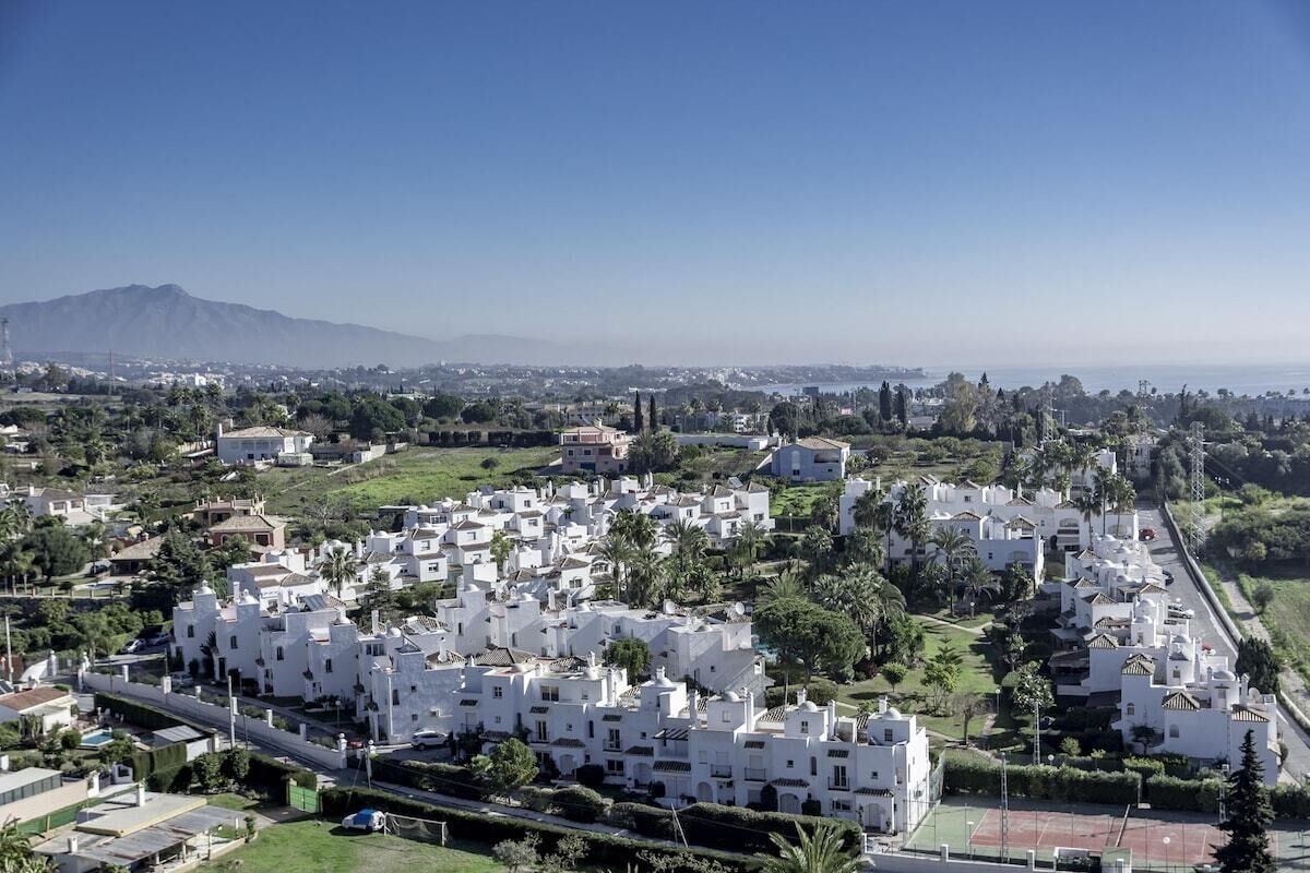 A residential area with white buildings, green spaces, and a mountain in the background.