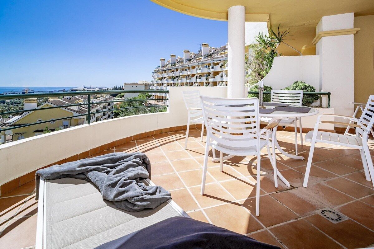 A balcony with a view of a coastal town, white chairs, a table, and a towel.