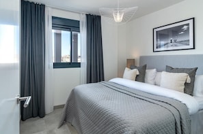 A modern hotel room with a large bed, grey bedding, a city view through the window, and a framed picture on the wall. - Inf3.6k - Luxury Apartment Perfect Location (Estepona)