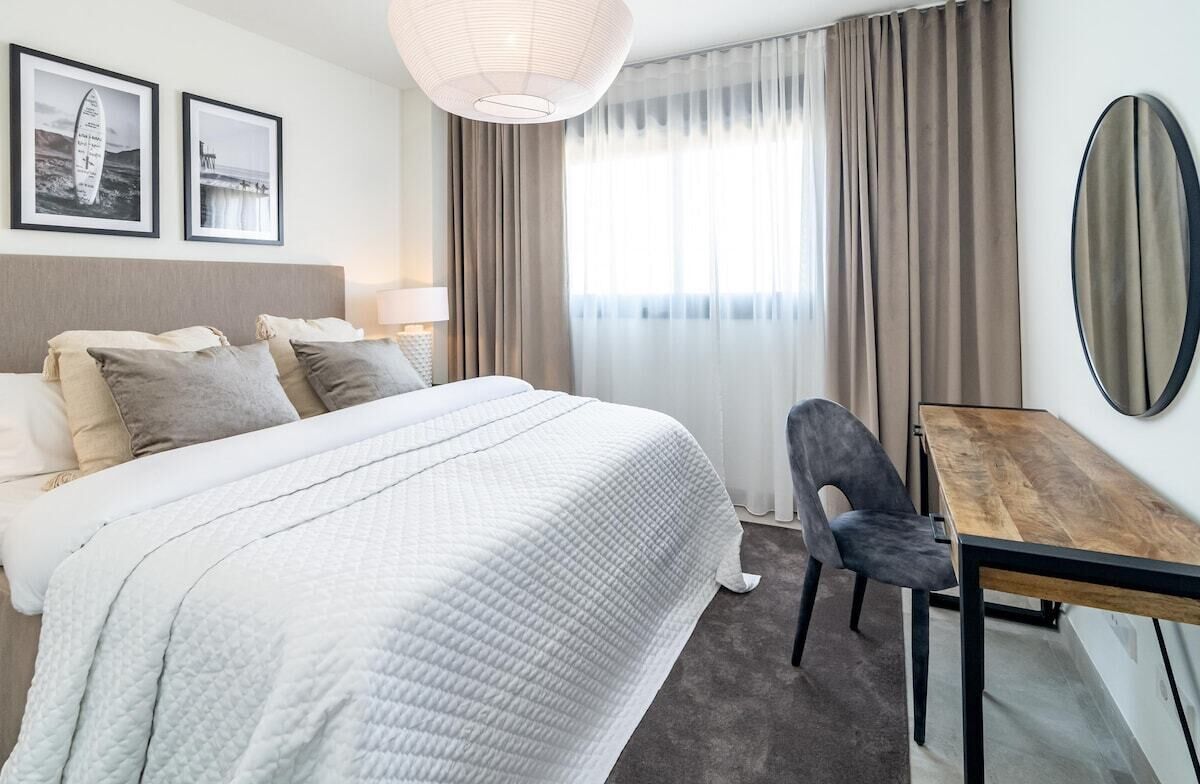 Een hotelkamer met een groot bed, een bureau en een stoel. Twee ingelijste foto’s hangen aan de muur, een groot raam met gordijnen en een ronde spiegel.