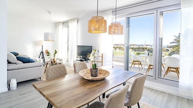 A modern living room with a wooden dining table, beige chairs, and a large window offering a view of the outdoors. - Lm12b- Exclusive 2 Bedroom Apartment in Le Mirage (Estepona)