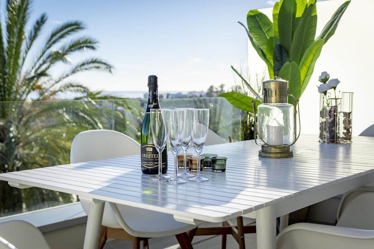 Ein Tisch gedeckt mit einer Weinflasche, Gläsern und einer Kerze auf einem Balkon mit Blick auf Palmen und einen klaren Himmel.