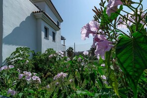 Um edifício com fachada branca e uma pequena janela, rodeado por vegetação exuberante e flores cor-de-rosa.