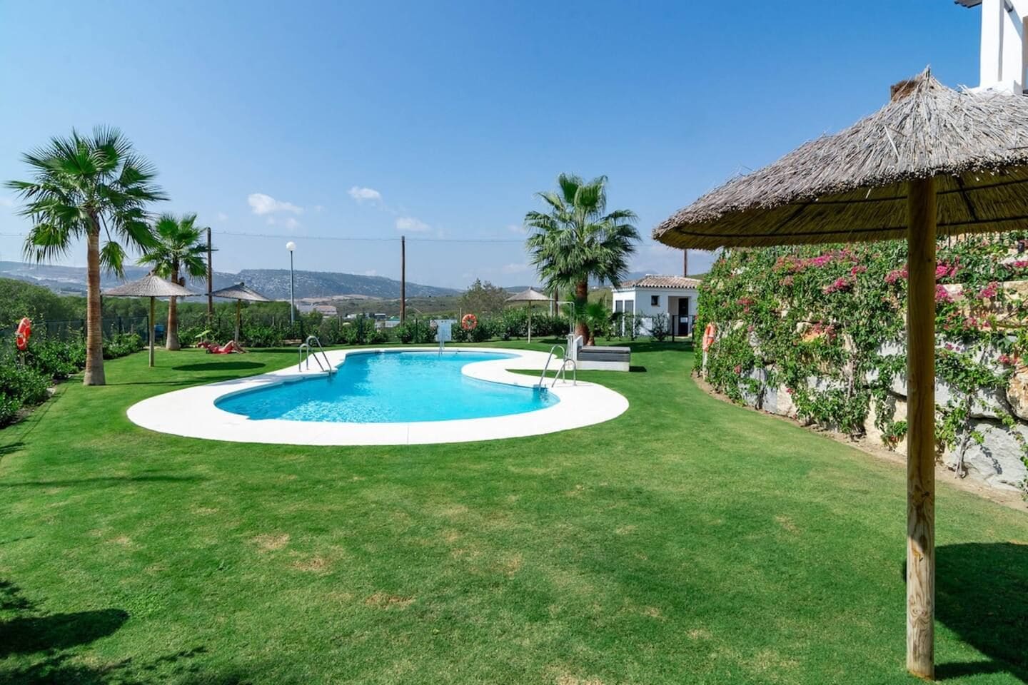 Una piscina con plataforma blanca, rodeada de césped cuidado, palmeras y una estructura de sombrilla de paja.