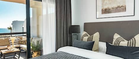 A hotel room with a bed, a nightstand, a lamp, a view of the sea, and a balcony with outdoor seating.
