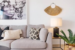 Ein modernes Wohnzimmer mit einem grauen Sofa, gemusterten Kissen, einer Wandlampe und einem dekorativen Wandbild.