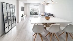 Un salon moderne avec un canapé blanc, une table à manger et quatre chaises, ainsi qu’une grande fenêtre donnant sur des immeubles.