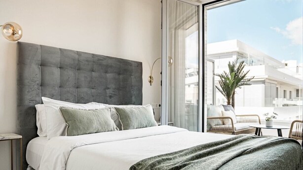Une chambre moderne avec un grand lit, une porte vitrée donnant sur un balcon et une vue sur les immeubles.